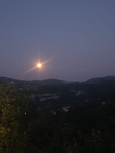 This image depicts a twilight landscape overlooking a valley near Fumane, Italy. The sky is a deep, hazy purple, with a bright, glowing full moon positioned slightly off-center to the left. The moon emits a prominent starburst-like flare. Below the sky, rolling hills covered in dark foliage stretch across the frame. In the middle ground, there appear to be scattered houses or structures nestled within the landscape, some emitting faint lights. A steep, rocky cliff face is visible in the mid-ground, suggesting a canyon or gorge. In the foreground, the dark silhouettes of leafy branches and foliage frame the lower portion of the image, adding depth and a sense of being on a vantage point. The overall atmosphere is serene and nocturnal.