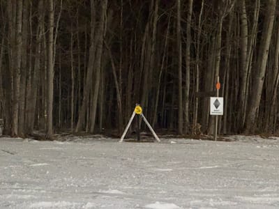 A snowy landscape at night with a dark forest as a backdrop. In the foreground, a trail closure sign is supported by white poles. To the right, a black diamond "most difficult" sign is visible. The ground is covered in snow with faint ski tracks. The forest consists of tall, bare trees, creating a stark contrast to the white snow.