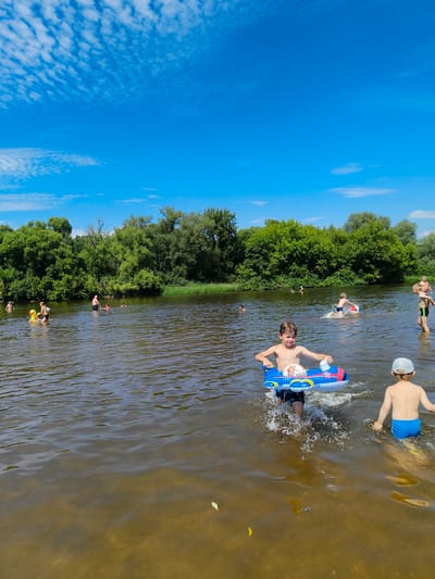 In Uspenskoye, Russia, on a sunny day with a bright blue sky dotted with wispy clouds, several individuals are enjoying a swim in a wide, calm river. The foreground features two young boys. One boy, in the center foreground, is partially submerged in the water, holding a blue and red inflatable toy shaped like an airplane with a soccer ball inside. He is splashing as he moves forward. To his right, another boy in blue swim trunks is also in the water, facing away from the camera.
Further into the river, other people can be seen. On the left, a child is playing with a yellow inflatable duck. Several other swimmers are visible further back, some appearing to be adults. The riverbank on the far side is densely lined with lush green trees and foliage. The water is murky, suggesting a natural river environment. The scene depicts a recreational gathering in a natural outdoor setting.