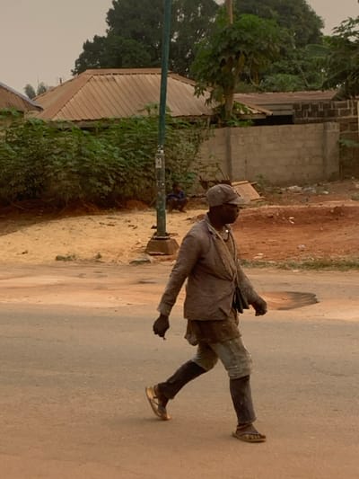 A man walks along a road in a rural setting. He wears dark clothing, including long sleeves, shorts, long socks, and shoes. A hat covers his head, and a bag hangs over his shoulder. In one hand, he holds a small, dark object. The road is paved, and buildings of brick and corrugated iron stand to the side, half-hidden by trees. A green metal pole stands near the edge of the road. A small child squats beside the pole.