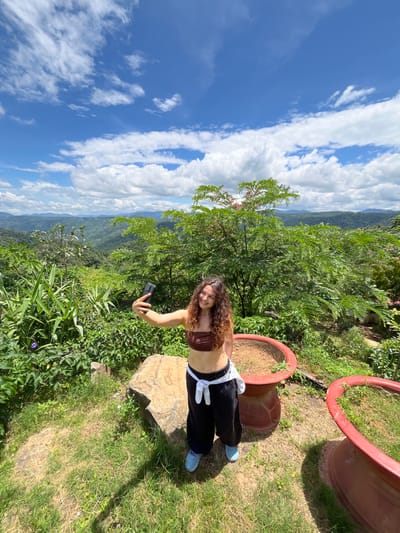 A young woman with curly brown hair and a brown tube top is taking a selfie with her phone. She is standing on a grassy area next to a large rock and two large, red planters. She is wearing black pants with a white shirt tied around her waist and blue sneakers. The background is a lush green landscape with rolling hills and a bright blue sky with white clouds. The scene is outdoors, possibly at a viewpoint in Sơn Điền, Vietnam, given the verdant, hilly terrain and tropical vegetation.