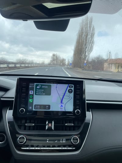 The image captures an interior view from a moving vehicle on a highway in Solak, Armenia, at approximately 13:21 under an overcast sky with visible moisture on the windshield.

The foreground is dominated by the vehicle's black dashboard and central infotainment system. The infotainment screen, displaying a navigation application (likely Apple CarPlay), shows the current time as 13:21 and an estimated arrival time of 13:42, indicating a 21-minute journey over 29 kilometers to "Yasaman Sevan's Restaurant." The vehicle's current speed is registered at 86 km/h, heading northeast (NE) on a road labeled "AH83." Below the screen, the vehicle's climate control panel indicates a temperature setting of "LO" with "SYNC" active. A red triangular hazard light button is centrally located between air vents.

Through the windshield, a multi-lane asphalt highway is visible, curving slightly to the right. A white corrugated barrier runs along the left side of the road. The right side of the highway is lined with tall, slender, bare deciduous trees. Further beyond the trees, a small, light-colored, single-story building with a dark roof is visible. Several blue road signs are partially visible on the right shoulder, beyond the trees. The sky is uniformly grey with heavy cloud cover, and small water droplets are evident on the windshield's exterior surface, suggesting rain or recent precipitation. No individuals are visible in the scene.