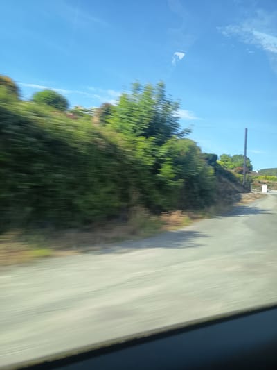 The image captures a motion-blurred view of a rural roadside in Killarney, Ireland. The scene is dominated by a lush green hillside covered in trees and vegetation on the left side. A paved road curves gently into the distance on the right. A tall utility pole stands near the roadside, and a small white structure, possibly a mailbox or a small building, is visible further down the road. The sky is a clear blue with scattered white clouds, suggesting a bright, sunny day. The motion blur indicates the photo was taken from a moving vehicle, likely a car.