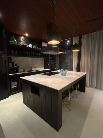 The image depicts a modern, unoccupied kitchen interior located in Batuan, Indonesia. The space features a dark wooden plank ceiling and dark wooden cabinetry throughout. A prominent central kitchen island with a light-colored marble countertop houses an integrated black four-burner gas stovetop. A light blue, wavy glass bowl and two small white rectangular objects are present on the island's surface. Three gold-finished metal bar stools with circular seats are tucked under the far side of the island. A rectangular recess is visible on the near side of the island's base.

Overhead, three dark, bell-shaped pendant lights with illuminated warm-toned inner surfaces hang from the ceiling, with two positioned directly above the island. A dark-paneled range hood with a metallic underside extends from the ceiling above the stovetop.

The back wall features a light-colored marble backsplash above dark wooden base cabinetry. A double sink with dark fixtures is visible. Various small appliances, including a coffee maker and toaster, are situated on the counter. Open dark wooden shelving above the main counter displays assorted glassware (wine glasses, tumblers) and decorative objects, including a large green wavy glass piece and several smaller teal-blue glass items.

To the left, a built-in black refrigerator and an integrated small beverage cooler are part of the cabinetry. On the far right, built-in oven units are visible. The right wall features a large window covered by sheer, floor-length white curtains. The floor is composed of large, light-toned square tiles. No individuals or active processes are present.
