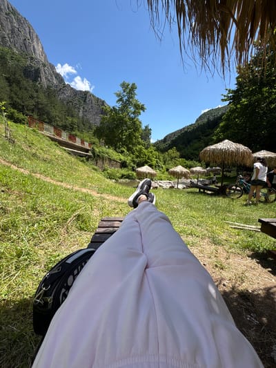 A first-person perspective shot captures a person relaxing on a wooden bench in a lush green park area. Their legs, clad in light pink pants, are extended forward, with their feet, wearing black and white sneakers, resting on the bench. A black bag with silver hardware is placed to the left of their legs.

The environment features a vibrant green lawn interspersed with patches of dry grass and scattered twigs. In the mid-ground, several thatched umbrella-like structures provide shade. A light blue bicycle stands near one of these umbrellas, with a person wearing a white t-shirt and denim shorts standing beside it. Another person, dressed in a dark shirt, is visible further back.

Beyond the park area, a shallow river flows, bordered by rocks and more greenery. A red and white railing runs along a raised path on a grassy slope to the left, which leads up to a sheer, rocky mountain face. The mountain is partly covered with dark green trees. The sky is a clear, bright blue with a few scattered white clouds. The scene is bathed in bright sunlight, suggesting it is a warm, sunny day. The location is identified as Zgorigrad, Bulgaria.