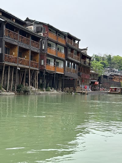 The image depicts a waterside scene in the city of Fenghuang, China. A multi-story building with a traditional dark wood and tile design is situated alongside a body of water, supported by wooden stilts. The building has multiple balconies and windows, with decorative wooden screens. To the right, the water's surface reflects the building. In the background, across the water, there are more buildings and green foliage. A boat carrying people is visible on the water. The sky is overcast.
