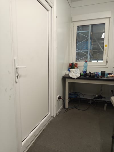 The image depicts the interior of a room, likely a portable office or storage unit, located in Vlkanová, Slovakia. A white door with a silver handle and lock is visible on the left side of the frame. To the right, a window looks out onto what appears to be a warehouse or industrial facility with tall shelving units. In front of the window is a dark grey table with various items placed on its surface, including a white cloth, a blue bottle of water, a pair of headphones, and a black cup. Below the table, an array of cables and electrical components are visible, suggesting a workspace or equipment area. The floor is a dark, speckled concrete or epoxy material. Electrical outlets are present on the wall near the door and the table. The overall impression is one of a functional, utilitarian space.