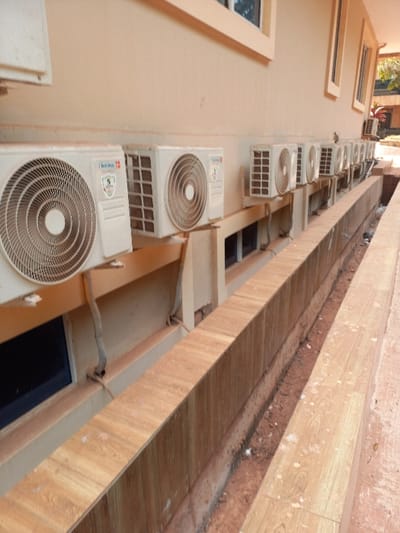 The image depicts the exterior wall of a building in Oturkpo, Nigeria, featuring a linear arrangement of approximately ten to twelve white outdoor air conditioning condenser units mounted above a series of rectangular windows. The building wall is painted a light cream color, and the windows have white frames. Beneath the air conditioning units and windows, a low, tiled barrier or planter box extends horizontally, clad in light brown wood-grain tiles. Adjacent to this barrier, a narrow trench filled with reddish-brown earth and minor debris runs parallel to the building. Electrical conduits and refrigerant lines are visible connecting the air conditioning units to the building wall. No individuals are present in the scene. The overall lighting suggests daylight conditions.