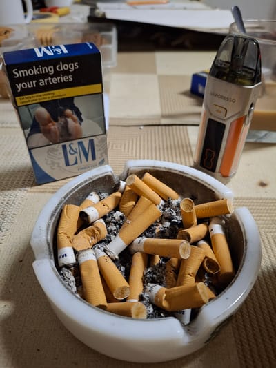 The image depicts a close-up scene featuring an ashtray filled with used cigarette butts and ash. Next to the ashtray is a pack of L&M Blue cigarettes with the health warning "Smoking clogs your arteries" prominently displayed. Adjacent to the cigarettes is a Vaporesso brand vape device. The background has a neutral, somewhat textured surface. The scene is not identifiable as being in Brunsbüttel, Germany, or any other specific location.