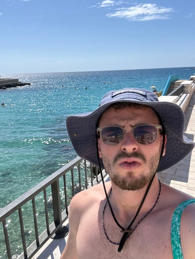 The image is a selfie captured by a male individual on a coastal promenade in Nardò, Italy. The subject, visible from the mid-chest upwards, wears a blue bucket hat with a white "Supreme" logo and light-framed sunglasses that reflect urban structures. He has a short beard, is bare-chested, and wears two necklaces: one beaded and one with a black cord and a brown, flat pendant. A teal and green patterned fabric is visible on his left shoulder. His facial expression features slightly pursed lips and subtly furrowed brows.

The scene is set under a clear blue sky with bright sunlight reflecting on the sparkling blue-green water. A black metal railing lines the promenade in the foreground. In the midground and background, the Ionian Sea contains several swimmers and one individual on a small watercraft near a rocky breakwater to the left. To the right, a stone promenade continues, featuring a distinct set of bright blue stairs descending to the water. Further along the coastline, a white umbrella is visible. This specific coastal configuration of the promenade, blue stairs, and rocky features is characteristic of the Santa Caterina di Nardò area.