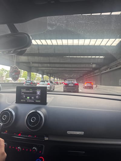 An interior view from a vehicle depicts a multi-lane highway scene under a concrete overhead structure in Villeneuve-la-Garenne, France, likely a covered section of the A86 autoroute near the Port de Gennevilliers or the A15 interchange.
The scene is observed through the front windshield of an Audi vehicle, indicated by the dashboard design, circular air vents with chrome surrounds, vehicle controls, and an "e-tron" badge on the passenger side. A partial view of a human hand is visible on the steering wheel, suggesting a driver is present. An infotainment screen in the center of the dashboard displays a phone interface with "Raccroch.", "Free", "Options", a phone number, "9:40", "Boîte voc. 18:03", and "Réglages" text. A rearview mirror hangs from the windshield, with a "CAR WASH" air freshener suspended below it. The windshield is smudged with dust and debris.
The highway ahead shows moderate to heavy traffic, with several passenger vehicles, including a dark SUV (possibly an Audi) and a white car, visible in multiple lanes. Their brake lights are illuminated, suggesting slow-moving traffic or a stop. The road is encompassed by a massive, multi-tiered concrete overhead structure. The ceiling of this structure features numerous parallel rows of rectangular light panels, providing artificial illumination to the roadway. To the far left, beyond the immediate concrete structure, green foliage and distant buildings are partially visible, with graffiti present on some concrete walls. The ambient light indicates daytime conditions, with illumination provided by overhead lights and filtered natural light.