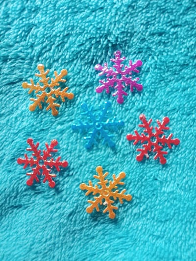 Six glittery snowflake decorations are arranged on a textured, turquoise fabric surface. There are two orange snowflakes, two red snowflakes, one blue snowflake, and one pink snowflake. The snowflakes are of varying sizes and are scattered across the fabric, not in any particular pattern. The fabric appears to be a soft, fuzzy material. There are no people or discernible actions taking place. The scene does not provide any information that could be used to determine its location in San Pascual, Philippines.