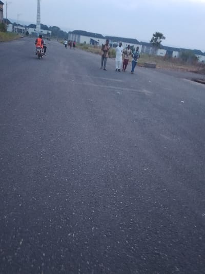 The image shows a paved road in Ogboko, Nigeria. A motorcyclist wearing an orange vest is driving away from the camera. Four men are walking along the side of the road, and a group of people are running in the distance. There are buildings visible in the background, and a tall tower is also present. The sky is overcast.