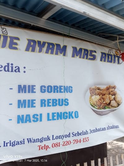 The image shows a sign for a food vendor in Kecamatan Anjatan, Indonesia. The sign is white with blue and yellow text. It reads "JE AYAM MRS ADIT" and lists the following menu items in blue text: "MIE GORENG," "MIE REBUS," and "NASI LENGKO." The sign also has a picture of a bowl of noodles with meat and vegetables. There is a green string tied around the top of the sign and a brown wooden fence behind it. The sign also includes the phrase "Irigasi Wanguk Lonyod Sebelah Jembatan alzaitun" and a phone number: "Telp. 081-220-794-155." The sign is mounted on a wall or other structure, and the top of the structure can be seen in the image. There are no people visible. The location is in Kecamatan Anjatan, Indonesia, as identified by the coordinates -6.42 latitude and 107.978 longitude.