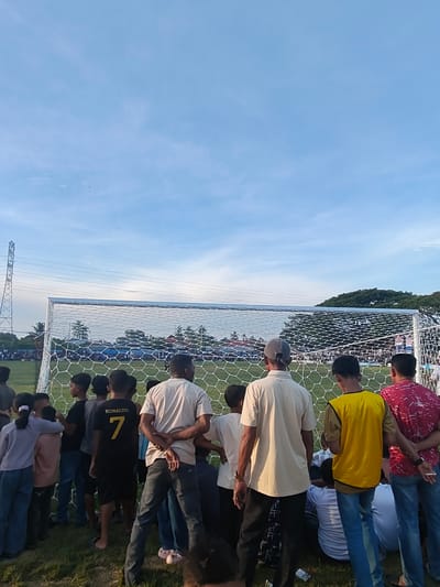 A crowd of people, primarily young males and some adult males and females, are gathered in front of a soccer goal net. The individuals are dressed in casual clothing, including t-shirts, jeans, and some wearing hats. Some are looking towards the soccer field, while others are facing away from the camera. The environment appears to be a soccer field with a large goal net in the foreground. In the background, there are trees, buildings, a power tower, and a vast sky with some clouds. The scene suggests an outdoor sporting event or gathering. The location is identified as Desa Meunasah Blang, Indonesia.