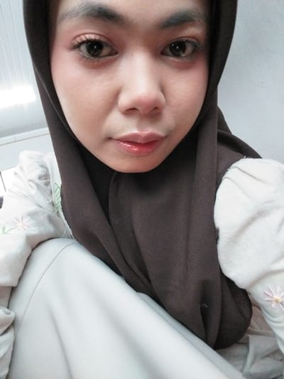A close-up, head-on selfie of a young Indonesian woman is captured from the chest up. She is wearing a dark brown hijab that covers her head and neck. Her face is framed by the hijab, with her eyes looking directly at the camera. She has light-colored skin and is wearing makeup, including mascara and lip gloss. Her clothing appears to be a light-colored garment with subtle floral embroidery on the sleeves. The background is a plain white wall, suggesting an indoor setting. The image is taken from a slightly low angle, looking up at the woman. The lighting is soft and diffused, highlighting her features. There are no visible geographical markers to indicate the specific location in Tibojong, Indonesia.