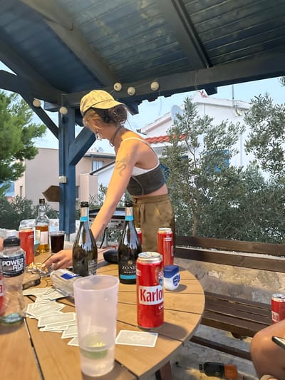 An outdoor scene captured at dusk or early evening in Betina, Croatia, shows a gathering centered around a wooden table beneath a dark wooden pergola adorned with string lights.
A female individual, viewed from the left and slightly behind, is the primary subject. She wears a beige baseball cap, a white bralette, a dark olive-green corset-style crop top, and pleated olive-brown trousers. Her hair is styled in a bun beneath the cap. A visible tattoo resembling a serpentine creature is on her right bicep. She is leaning forward, with her right hand extended towards items on the table.
The round wooden table in the foreground holds various items. From left to right: a clear plastic bottle labeled "POWERADE," a red can (partially obscured), a brown glass bottle (possibly spirits) with a dark liquid in a clear glass next to it, an ashtray containing multiple cigarette butts, and a clear plastic bag. Two light blue-labeled bottles of "ZONIN PROSECCO" stand prominently. Multiple red cans labeled "Karlovačko Lager" are visible, with at least three clearly identifiable. A small blue and white container, resembling Vaseline, is also on the table. Playing cards are scattered across the table surface. In the immediate foreground, a translucent plastic cup with a lime wedge at the bottom is visible.
In the background, a silver-colored outdoor grill is positioned behind the individual. Beyond this, dense green foliage, including a tall pine tree on the left and olive trees in the center, partially obscures white-plastered buildings with terracotta roofs, typical of Mediterranean architecture. A satellite dish is affixed to one of the distant rooftops. Below the table, a stone wall is visible, and a dark, cylindrical object rests on the ground.