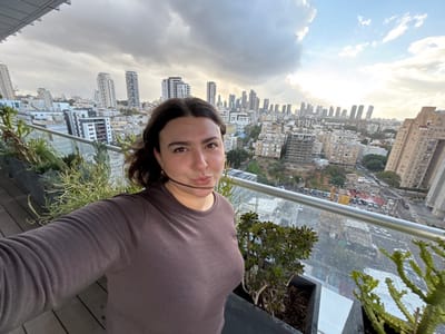 A female individual is depicted in a selfie, positioned on an elevated outdoor balcony or terrace in Bnei Brak, Israel. She has dark hair and wears a dark brown long-sleeved top. Her face is directed towards the camera, with her lips pursed in an expressive manner; a thin, dark strand of material or hair is horizontally across her mouth area. The foreground features a glass railing with a metallic top bar, behind which several potted plants, including succulents, are arranged. The background reveals a dense urban cityscape, characteristic of Bnei Brak, with a broad view extending towards the skyline of Tel Aviv, identifiable by numerous modern high-rise buildings and skyscrapers in the distance. Mid-ground shows various multi-story residential and commercial buildings, some appearing to be under construction, alongside streets with visible vehicular traffic. The sky is partially overcast with grey clouds, but areas of sunlight are visible towards the horizon, suggesting late afternoon or early evening.