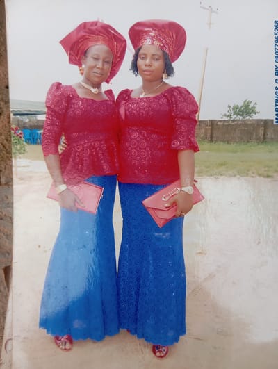 Two women stand side-by-side outdoors in what appears to be the city of Karsana 2, Nigeria. Both women are wearing matching traditional Nigerian attire. They have on red, intricately patterned blouses with puffy sleeves and matching headwraps. Their bottoms are long, flowing blue skirts made of a similar patterned lace fabric. Both women are holding red clutch purses. The woman on the left is wearing gold earrings and a gold necklace. The woman on the right has gold earrings and a gold necklace. The ground beneath them is a light-colored, dusty surface. In the background, a brick wall and some greenery can be seen. There is also a light pole and what looks like part of a tent or canopy structure to the left. A phone number is visible printed vertically on the right side of the image.