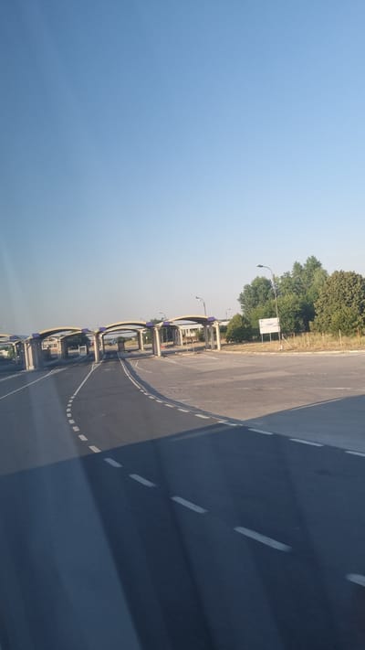 The image captures a view of the Giurgiu–Ruse Border Checkpoint in Giurgiu, Romania, on a clear day. The foreground shows a paved road with dashed white lane markings, curving slightly to the left. Shadows of an unseen object, possibly a vehicle, fall across the road. In the mid-ground, several arched structures of the border checkpoint are visible, with concrete pillars supporting yellow-canopied roofs. These structures appear to span multiple lanes of traffic. Beyond the checkpoint, trees and vegetation line the right side of the road. A white signpost is also visible in the mid-ground on the right. The sky is a clear, pale blue, indicating fair weather. The scene depicts an infrastructure point for border crossings, with no people or vehicles immediately apparent in the captured frame.