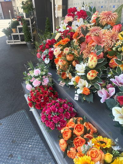 This media file features a vibrant display of artificial flowers arranged on tiered shelves outside a shop. The flowers are in various stages of bloom and come in a wide array of colors, including shades of orange, pink, red, yellow, white, and purple. Prominent among them are large, multi-petaled blooms resembling chrysanthemums and dahlias, alongside clusters of roses and smaller accent flowers. Some of the flowers are presented in bouquets, while others are individually displayed.
The setting appears to be an outdoor market or a florist shop's storefront in Clermont-Ferrand, France. The ground is paved with asphalt, and a diamond-plate metal cover is visible in the lower right portion of the image, possibly an access panel. In the background, there are glimpses of a building and some greenery.
The arrangement of the flowers suggests they are for sale. There are price tags visible on some of the floral displays, with handwritten numbers and labels, though the text is not fully legible. One tag clearly shows "Fleurs" (French for flowers).
The lighting suggests it is daytime, and the weather seems pleasant, with no indication of rain or harsh sunlight. The overall impression is one of abundance and visual appeal, highlighting the variety and artistry of the artificial floral arrangements. The focus is solely on the inanimate objects, with no people or animals present.