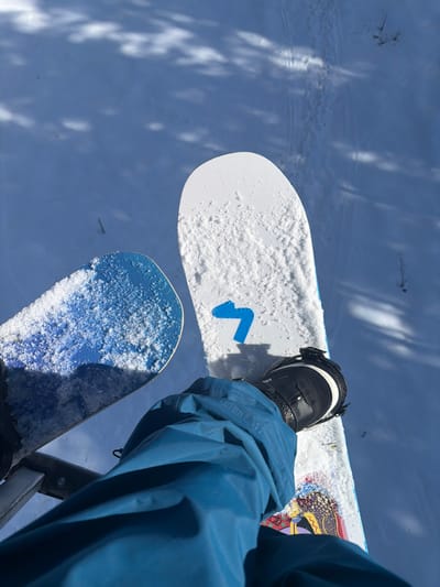 An elevated, first-person perspective photograph captures a winter scene in Lunnaya Polyana, Russia. The lower half of a person's body is visible, wearing blue snow pants and a black and white snowboard boot. The boot is secured into a binding on a white snowboard, which features a blue arrow-shaped graphic and is partially covered in granular snow. To the left, a second snowboard, blue and also partially snow-covered, is visible. Both snowboards are suspended, indicating the individual is likely on a chairlift or similar aerial transport mechanism, with a metallic support structure partially visible in the lower-left corner. Below, the ground is covered in undisturbed white snow, marked by visible tracks, potentially from skis or snowboards. Scattered tree shadows are cast across the snow, alongside sparse, dry vegetation protruding from the surface. The sky is not visible.