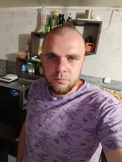 A man with a shaved head and a beard is standing in what appears to be a kitchen. He is wearing a purple t-shirt with a repeating pattern of cartoonish figures surfing. Behind him, there are shelves with various items, including bottles, jars, and what looks like a hookah. To his left, there is a microwave and a digital scale. The overall environment suggests a domestic setting. The image was taken in the city of Pishchane, Ukraine.