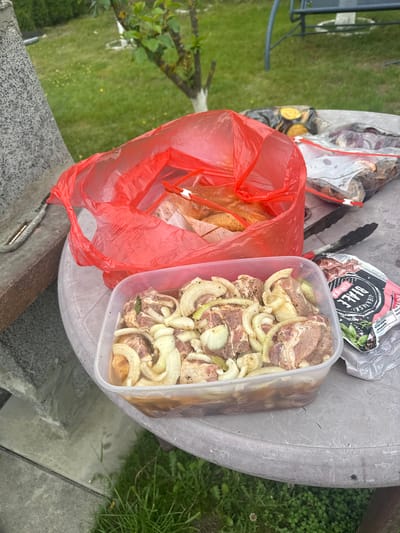 The image shows a collection of items on a round, concrete table in an outdoor setting, possibly a backyard or patio. A clear plastic container holds marinated meat, intermingled with sliced onions. Next to it is a red plastic shopping bag containing skewers with roasted food. Behind the container is a package of "Kielbaski Biale" sausage, a pair of tongs, and another package. The location is most likely in Lipnik, Poland, given the packaging of the sausages. The surrounding environment includes greenery, suggesting a garden or lawn. The overall scene implies preparation for a barbecue or outdoor meal.