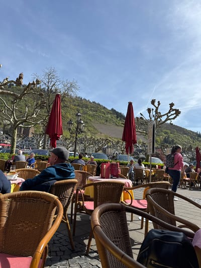 The image shows an outdoor cafe scene in Cochem, Germany. Wicker chairs are prominent in the foreground, arranged around tables, some with red tablecloths and menus. People are seated at the tables. A man in a blue jacket and cap is visible at one table. In the background, there are views of the town, with trees, buildings, and a hillside. Red umbrellas are positioned among the tables, and the sky is blue with a faint contrail.