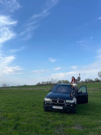 The image depicts a black BMW X5, license plate CD 2122 CX, parked in a green field under a bright blue sky with scattered clouds. A woman with red hair is on top of the car. Another person is near her. The car's driver-side door is open. The location appears to be in a rural area near Barkach, Bulgaria.