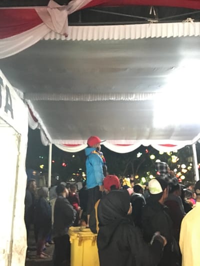 A group of people are gathered under a white tent, likely in a public square, located in the city of Kecamatan Batu, Indonesia at coordinates -7.867, 112.514. There are several people standing around a yellow garbage bin. A young boy wearing a blue jacket and red hat is standing on top of the bin. The environment is dim, as the scene appears to be taking place in the evening. The tent is decorated with red and white curtains. There is a slight red and white flag hanging from the top of the tent. A man in a plaid shirt is standing behind the boy. A woman in a black hijab is standing next to the man in a plaid shirt. The boy is looking up at something above him. Several other people are standing behind the group of people in front of the garbage bin. It is dark outside of the tent and there is a line of streetlights illuminating the trees and other foliage. This appears to be a scene from a community gathering in Kecamatan Batu.