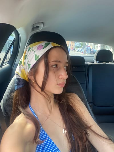 A young woman with long, dark hair and a colorful headscarf is sitting in the back seat of a car. She is wearing a blue and white gingham bikini top and a delicate necklace with a "W" pendant. Her hair is partially covering her face as she looks out the window. The interior of the car is visible, with the dark upholstery of the seats and the headrests. Through the car window, a street scene in Torbole, Italy can be seen. A gas station with visible prices and several people are present. Buildings and a street are also visible in the background. The lighting suggests it is daytime.