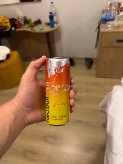 A person holds a can of Red Bull The Apple Edition. The can is primarily red and yellow. The background shows a room with a bed, wooden floors, a water bottle, and a wooden cabinet. The exact location within Tepebaşı, Turkey cannot be determined from the image.