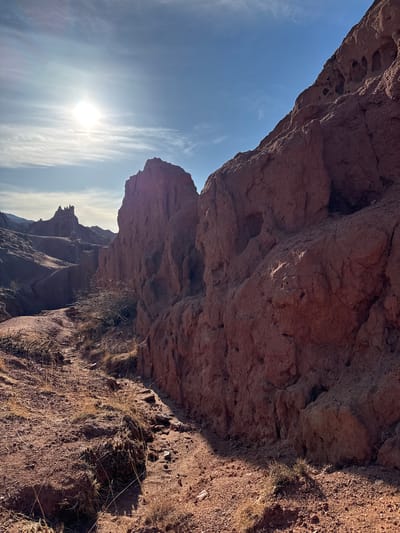 The image presents a rugged, sunlit landscape in Tosor, Kyrgyzstan, dominated by imposing reddish-brown rock formations under a bright, partly cloudy sky. The scene is oriented vertically, showing vast rock faces rising towards the sky from the left and right, framing a view into a canyon or valley.
The primary subjects are the large, weathered sandstone or conglomerate rock formations, exhibiting rough, eroded textures, deep shadows, and sunlit surfaces. In the foreground, particularly on the right, the terrain consists of reddish-brown soil and gravel, sparsely covered with dry, yellowish-brown grasses and small, tough shrubs. These foreground elements cast distinct shadows, indicating strong, direct sunlight.
The sky occupies a significant portion of the upper left, displaying a vibrant blue hue with wispy, streaky white clouds. The sun is prominently featured in the upper middle-left, appearing as a bright, glowing orb with noticeable lens flare, suggesting either early morning or late afternoon. Its low position in the sky creates dramatic lighting, highlighting the contours of the rocks and casting long shadows.
In the mid-ground and background, further distinct rock formations are visible, receding into the distance and appearing somewhat hazy, adding depth to the scene. These distant peaks and eroded structures contribute to the grandeur of the natural setting. No human activity or identifiable objects are present, emphasizing the untouched wilderness. The weather is clear and sunny. No text is visible in the image. The overall impression is one of natural majesty and rugged beauty, characteristic of arid or semi-arid mountain landscapes.