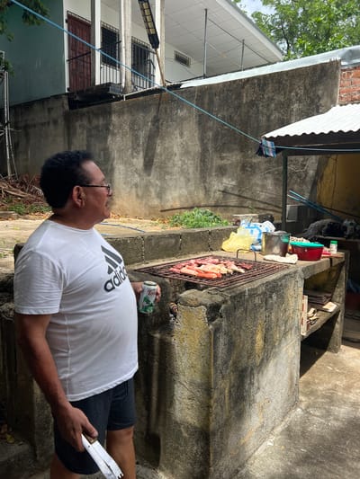 A man wearing a white Adidas t-shirt and dark shorts stands in front of an outdoor grill in what appears to be a residential backyard in San Juan del Sur, Nicaragua. He is holding a pair of metal tongs and a can of beer in his left hand. The grill is laden with various cuts of raw meat, including sausages and what look like chicken pieces. Behind the grill, a concrete structure with shelves holds plastic bags, a metal pot, a red container, and cleaning supplies. The environment includes a textured concrete wall in the background, some overgrown vegetation, and parts of a building with a metal roof. The lighting suggests it is daytime, with shadows cast on the ground.