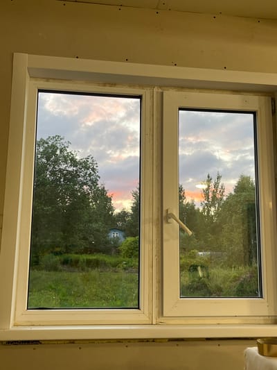 The image shows a double-paned window with white PVC frames, looking out onto a landscape at dusk. The sky is filled with dramatic clouds, colored with hues of pink, orange, and gray, indicating either sunset or sunrise.
Through the left pane, a dense canopy of green trees dominates the view. In the distance, nestled among the trees, is a small, light blue building with a white roof and a visible white window. The foreground on this side consists of a grassy area.
The right pane offers a similar view of lush green trees and foliage. The sky here is also cloudy and tinged with sunset colors. A white window handle is visible on the frame of the right pane.
The interior of the room from which the photograph is taken is partially visible. The wall appears to be unfinished, with visible screws and what looks like drywall. A white window sill extends below the window. A golden bowl and a white cloth are visible on a surface in the lower right corner of the image, suggesting this is an indoor scene.
The overall impression is one of a rural or semi-rural setting, viewed from inside a building under construction or renovation. The location, Клобушнево, Russia, suggests a potentially temperate climate, consistent with the visible trees and the appearance of the sky.