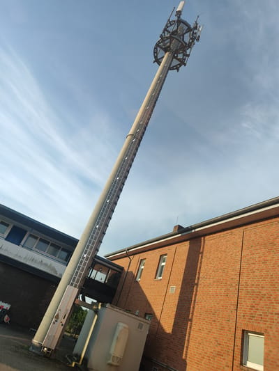 A tall, grey cell tower is the central subject of this image, tilted diagonally from the bottom left to the top right. The tower has a wide base and narrows towards the top, where it supports a circular platform with antennas. A ladder runs vertically along the side of the tower.
To the left of the cell tower, there is a low-rise building with a dark brick base and a lighter upper section featuring windows and blue accents.
To the right of the cell tower is a brick building with a prominent red brick facade. This building has several windows, some of which are dark. A shadow from the cell tower stretches across the brick wall.
At the base of the cell tower, there is a white, box-like structure with vents, possibly housing equipment. The ground in the foreground appears to be asphalt or gravel.
The sky is blue with wispy white clouds, suggesting daylight. The scene is outdoors, likely in an industrial or commercial area of Oldenburg in Holstein, Germany, given the architecture and the presence of a cell tower. There are no people visible in the image.