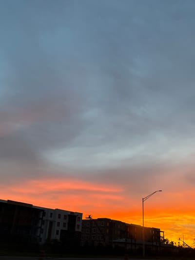 A vibrant sunset illuminates the sky over apartment buildings under construction in North Port, Florida. The sky transitions from a soft blue at the top to fiery oranges and yellows near the horizon. Silhouetted against the colorful sky are several multi-story apartment buildings, some appearing partially completed with exposed framing and scaffolding. A streetlamp stands prominently in the foreground, its arm extending upwards and to the right. The lower portion of the image is dark, obscuring the ground and any immediate surroundings. The scene captures a moment of dramatic natural beauty juxtaposed with urban development.