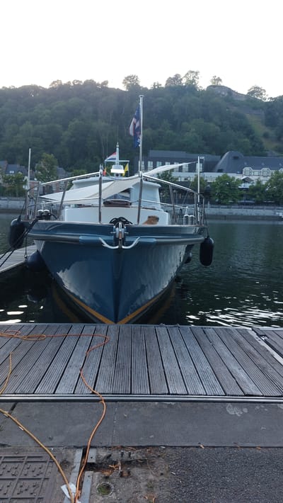 This image depicts a boat docked alongside a wooden pier. The boat, a dark blue vessel with a yellow trim, has its bow facing the viewer. Several flags are visible on the boat's mast, including what appears to be a flag with stars and stripes and another flag with red and white stripes. The boat is tied to the pier with ropes, and black fenders are attached to its side.

The pier is constructed of weathered gray wooden planks and runs along the right side of the image. Beyond the pier and the boat, a body of water is visible, reflecting the surrounding environment. In the background, a steep, tree-covered hill rises, with buildings nestled amongst the trees. The architecture of these buildings suggests a European setting, with a prominent tiled roof visible. The sky on the left side of the image is bright and overcast, indicating either early morning or late afternoon. The overall scene suggests a peaceful, still atmosphere. There is no visible text in the image.