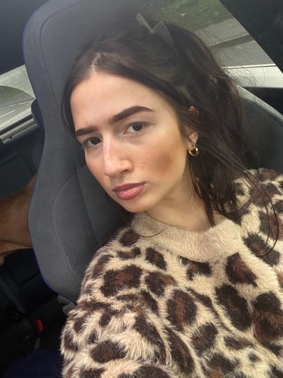 A young adult female is depicted in a close-up portrait, seated inside a vehicle in Broxbourne, United Kingdom. She has dark brown hair pulled back in a high ponytail secured with a light-colored claw clip, and is wearing a fuzzy, leopard-print sweater in shades of beige, brown, and black. A gold-colored hoop earring is visible on her right ear. She looks directly towards the camera with a neutral expression. To her left, partially obscured by a gray car seat with blue trim, is a reddish-brown animal, likely a dog. The blurred background outside the vehicle's window indicates a road with a white line and some vegetation.