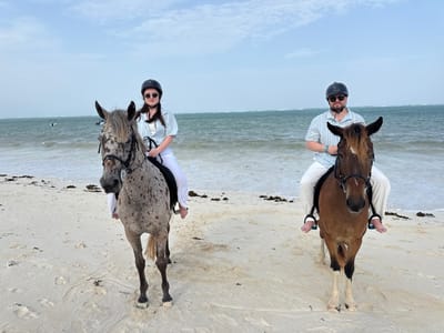 Two individuals on horseback are positioned on a sandy beach at Kiwengwa, Tanzania, with the Indian Ocean in the background under an overcast sky. The person on the left, a female, wears a black riding helmet, dark sunglasses, a light blue striped short-sleeved shirt, and white pants, riding a dapple grey horse with dark spots. The person on the right, a male, also wears a black riding helmet, dark sunglasses, a light blue striped short-sleeved shirt, and white pants, with a blue wristband on his left wrist, riding a bay horse with a flaxen mane and blaze. Both riders are barefoot, with their feet in the stirrups. The horses are fitted with dark bridles and stand stationary on the light-colored sand, which contains scattered dark seaweed near the waterline. The ocean exhibits gentle waves, and several dark objects, possibly small vessels or buoys, are visible further out on the water.