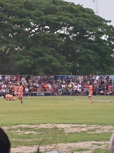 A soccer match is underway with spectators in the background. The field is mostly green grass with some bare patches. Three soccer players in orange and white uniforms are visible on the field. One player is kneeling on the ground, another is standing and looking forward, and a third is walking away from the camera. In the background, a large crowd of spectators sits and stands behind a blue fence. The spectators are wearing a variety of clothing in different colors. Behind the spectators, a large, leafy green tree dominates the upper portion of the frame. A white cell tower is visible in the upper right corner of the image. The lighting suggests it is daytime, likely late afternoon or early evening, with overcast skies. The scene appears to be an outdoor sporting event in a rural or semi-rural location. A black sign with white text reading "22 STORE" is visible on the field behind the spectators.