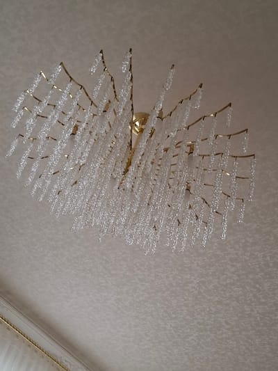 A chandelier hangs from a textured, light-colored ceiling. The chandelier has a gold-colored frame with numerous strings of clear, faceted beads that hang down in various lengths, resembling icicles or fringe. One light bulb is visible within the chandelier structure. The ceiling has a subtle, repeating pattern. In the lower left of the image, a portion of a window treatment with decorative gold trim is visible. There is no information in the image to indicate its location in Urus-Martan, Russia.