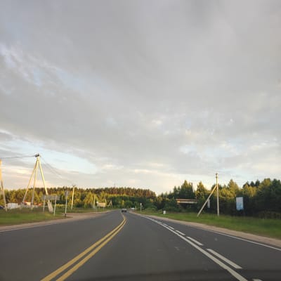 The image captures a rural road scene at dusk, with a wide asphalt road curving to the left. The road is marked with a double yellow line in the center and a white dashed line on the right shoulder. On the left side of the road, there are power lines supported by tall, triangular wooden poles, and a small collection of parked vehicles including a white van with red flashing lights, and what appears to be construction equipment. Beyond the road and to the left and center, a dense forest of green trees forms a backdrop, with a clear sky with wispy clouds above. On the right side of the road, there is a sign with the Cyrillic text "БЕТОН 40107" and another blue sign with text, all set against the forest. A dark sedan and a lighter colored car are visible driving on the road in the distance. The overall impression is one of a peaceful, somewhat isolated, rural setting.