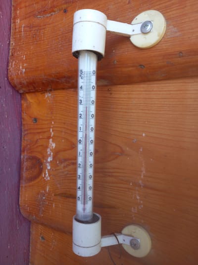 A close-up, high-angle, vertical shot shows a thermometer attached to a wooden wall. The thermometer is white with a glass tube containing a red liquid. The temperature scale is marked in Celsius, with markings for 0, 5, 10, 15, 20, 25, 30, and 35 degrees. The red liquid level is just below the 5-degree mark, indicating a temperature slightly above 4 degrees Celsius. The thermometer is held in place by white plastic brackets that are screwed into the wood. The wooden wall is made of rough-hewn planks with visible grain. The wall appears to be weathered and has some white splotches and dirt. The background is a blurred continuation of the wooden wall, with a hint of purple on the far left edge. The location is Adutiškis, Lithuania.