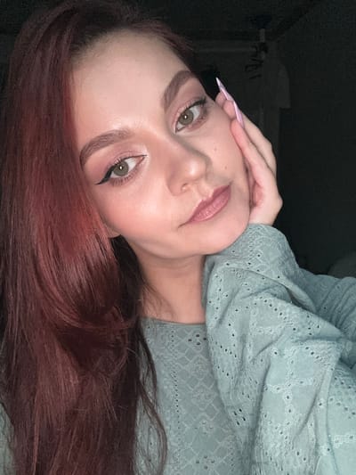 A close-up, rotated portrait shot of a woman with long, maroon hair. She's wearing light eye makeup with winged eyeliner and has pale pink lipstick. Her hand is near her forehead, and she's wearing a light green top with a lace pattern. The background is dark. This image was not taken in Buk-myeon, South Korea.