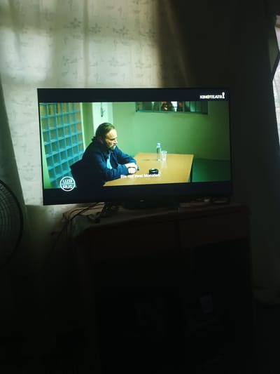 A television screen displays a scene from what appears to be a movie or TV show. The on-screen image shows a middle-aged man with a beard sitting at a wooden table in a room with light green walls and a wall of blue glass blocks. A bottle of water and a glass are on the table, along with an ashtray. Subtitles in yellow read "Bis vor zwei Monaten." In the bottom left corner of the screen, there is a circular logo with "UZB RUS" written inside. The TV is positioned on a dark wooden furniture piece. The background of the room, outside of the TV screen, is dark and indistinct, with a faint pattern on the wall behind the television. A round fan is partially visible on the left side of the frame. The overall lighting suggests it is nighttime or the room is dimly lit.