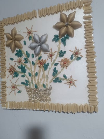 The image displays a wall-mounted art piece crafted from pasta. The central design features a bouquet of flowers, with larger, metallic-painted shell pasta flowers in gold and silver hues, and smaller, tan-colored shell pasta flowers with red bead centers. The stems and leaves are constructed from green and tan-colored pasta pieces. The entire artwork is framed with penne pasta arranged in a zig-zag pattern. The background is a plain white surface, and the artwork is mounted on a wall, possibly in a home or public space within the village of Бәйдібек би ауылы, Kazakhstan.