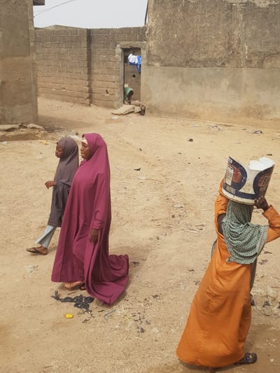The image depicts a street scene in Dutsinma A, Nigeria. Two women in traditional Islamic dress, one in a dark rose robe and the other in a grey robe and jeans, walk together on a sandy surface. A third woman, in an orange robe, walks in the opposite direction with a printed container on her head. Debris and trash are scattered on the ground. A partially-constructed brick building is on the left, with a figure visible in a window opening along with some hanging clothing.
