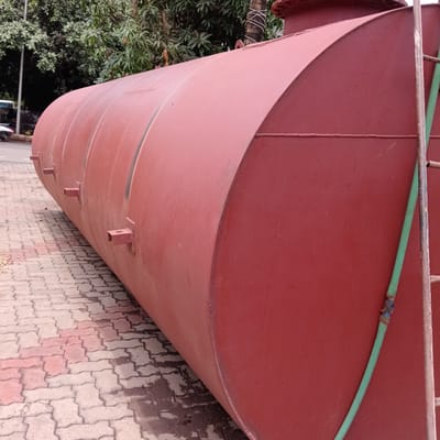 A large, cylindrical, maroon-painted metal tank rests on a paved surface of interlocking bricks in an outdoor setting in Marna, India. The tank has a rounded end visible in the foreground, with several square brackets welded along its length. A green hose is draped along the right side of the tank. A metal ladder rests against the right end of the tank. The background includes a street with vehicles and green foliage. The image appears to be taken at ground level, providing a close-up view of the tank and its immediate surroundings.